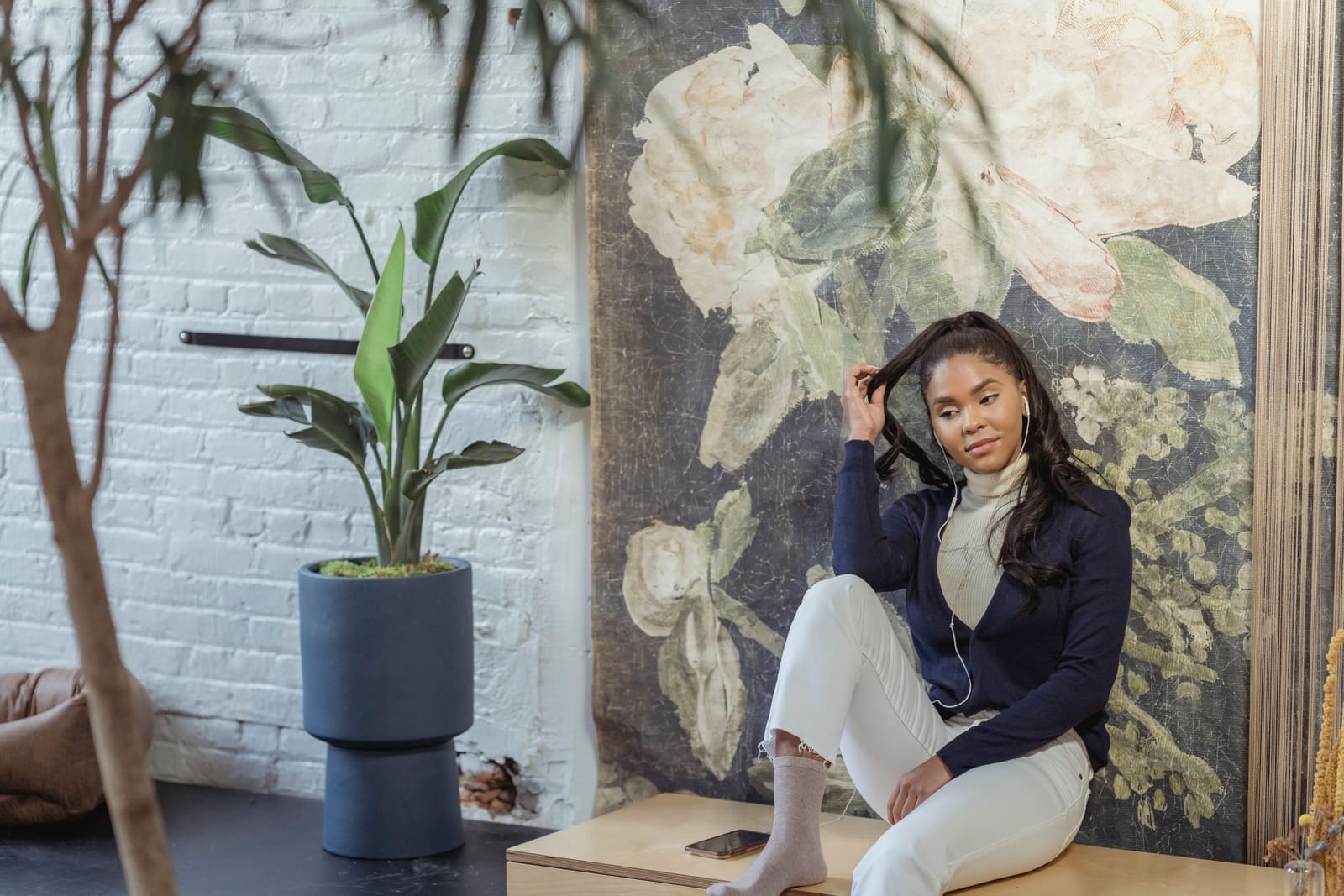 Black woman relaxing at home and listening to music through earphones in a bright room with plants and artwork.