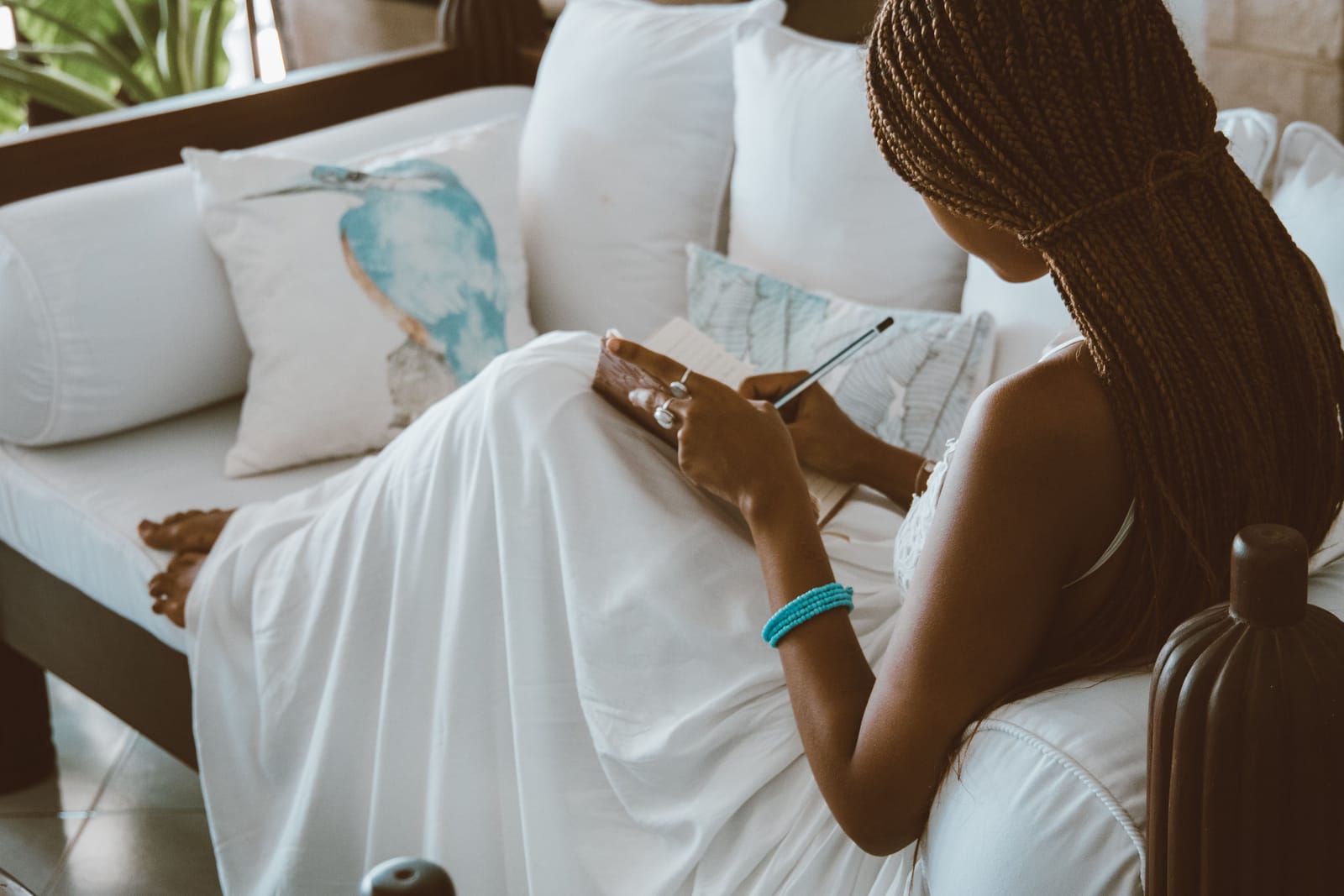 A Black woman in a white dress writing in a notebook while seated on a softly lit sofa at home.